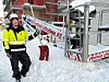 Mann med hjelm, gul signaljakke og snøskuffe på en byggeplass med mye snø.