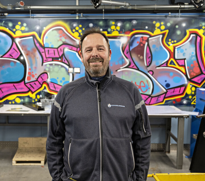 Sven Erik Knutsen er leder i NM-komiteen for blikk. Mann med skjegg i svart jakke foran verktøybenk der bakveggen med tagget med Blikk1 i friske farger.