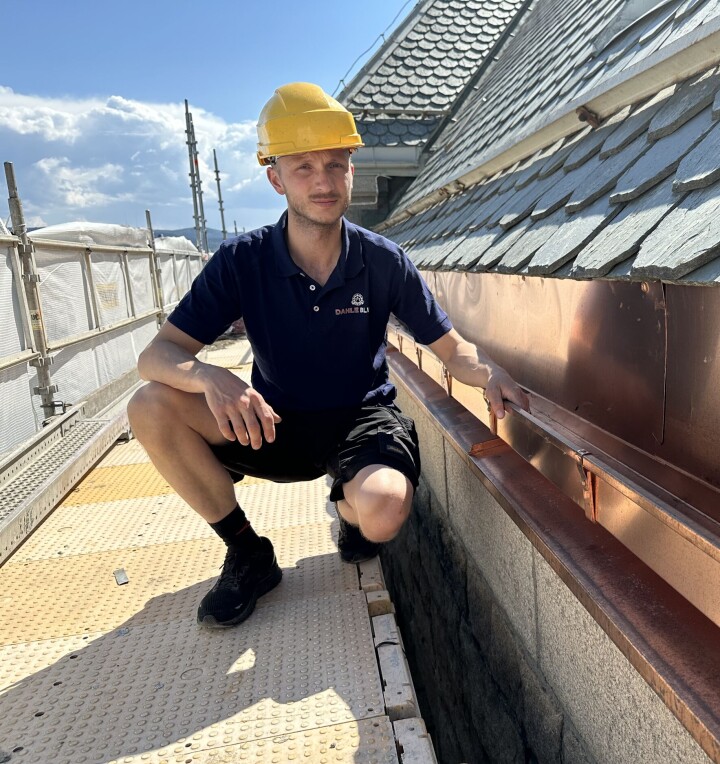 NTNU-bygget har fått nye håndlagede takrenne i kobber som skal vare i nye 100 år. Mann med gul hjelp og blå T-skjorte på stillas som viser frem en takrenne på taket.