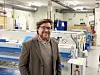 Person med briller og jakke står smilende i et moderne verksted med store maskiner.