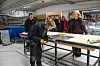Lærling kutter metallplate ved arbeidsbenk mens besøkende ser på i verksted