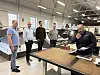 Fire menn i en verkstedhall ser på en arbeidsbenk med materialer og dokumenter.