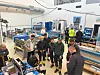 Personer står rundt en metallkonstruksjon i en lys industrihall med maskiner og arbeidsbenker.