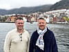 To menn står foran en havn med fargerike hus, vann og fjell i bakgrunnen.