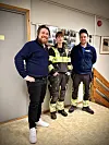 Tre personer står sammen innendørs, én i gul refleksbekledning og to i mørke klær, ved en trapp.