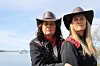 To kvinner i svarte klær og cowboyhatter står ved sjøen med en kystlinje bak.