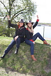 To kvinner i svarte hatter sitter på en gresskledd høyde ved vannet og vinker mot kamera.