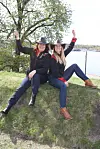 To kvinner i svarte hatter sitter på en gresskledd høyde ved vannet og vinker mot kamera.