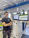 Smilende ansatt står ved en industrimaskin med to skjermer i en lys fabrikkhall.