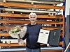 Mann står foran lagerreoler og holder en blomsterbukett og et innrammet diplom.