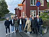 Flere personer står samlet foran en rød bygning med vinduer, skilt og blomster langs veggen.