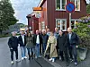 Flere personer står samlet foran en rød bygning med vinduer, skilt og blomster langs veggen.