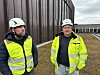 To arbeidere i verneklær og hjelm står utenfor et industribygg.