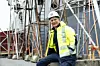 Arbeidstaker med hjelm og refleksjakke foran stillaser og stålkonstruksjoner på en byggeplass.