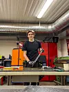Ung mann i svart T-skjorte står ved en arbeidsbenk i et verksted med verktøy og industrimaskiner.