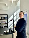 Person ved skrivebord i lyst kontor med hyller, lampe og sollys som faller inn gjennom vinduet.