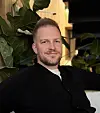Portrett av en smilende mann i svart genser foran grønne stueplanter.
