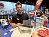 Mann ved en messestand holder metallutstyr, omgitt av produktprøver, skilt og en liten serveringsmodell.