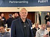 Mann i mørk jakke på Flexit-partnersamling med deltakere i bakgrunnen.